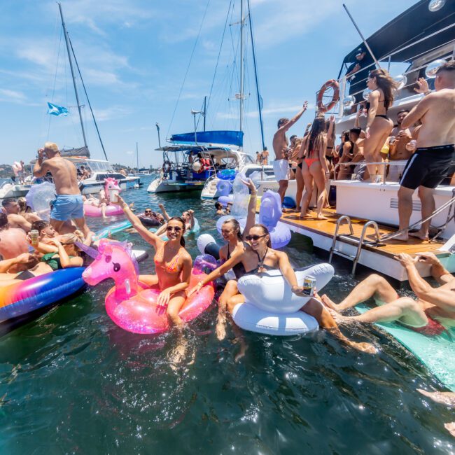 A lively boat party with people on inflatable rafts and floaties in the water. Several boats are docked nearby, and participants are enjoying the sun, swimming, and socializing. The atmosphere is festive and relaxed.