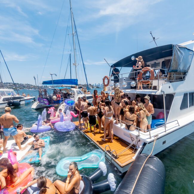 A group of people having a lively party on a yacht surrounded by inflatable floats in the water. Several boats are nearby under a sunny sky. Everyone is in swimwear, enjoying the festive atmosphere.