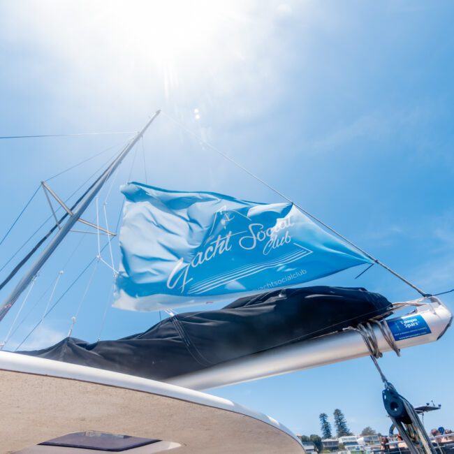 A blue flag with "Yacht Social Club" flutters atop a yacht under a bright sunlit sky. The yacht is docked near a coastal area with buildings and greenery in the background. A person is partially visible on the boat.