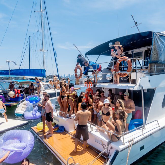 A lively party on a yacht with a group of people enjoying the sun and water. Some are on the deck, while others relax in inflatable floats in the water. The sky is clear and blue, and the scene is festive and vibrant.