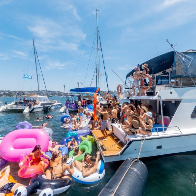 A lively gathering on a yacht with numerous people dancing and socializing. Many colorful inflatable floats, including unicorns and swans, fill the water around the boats. The sky is clear, suggesting a sunny day.