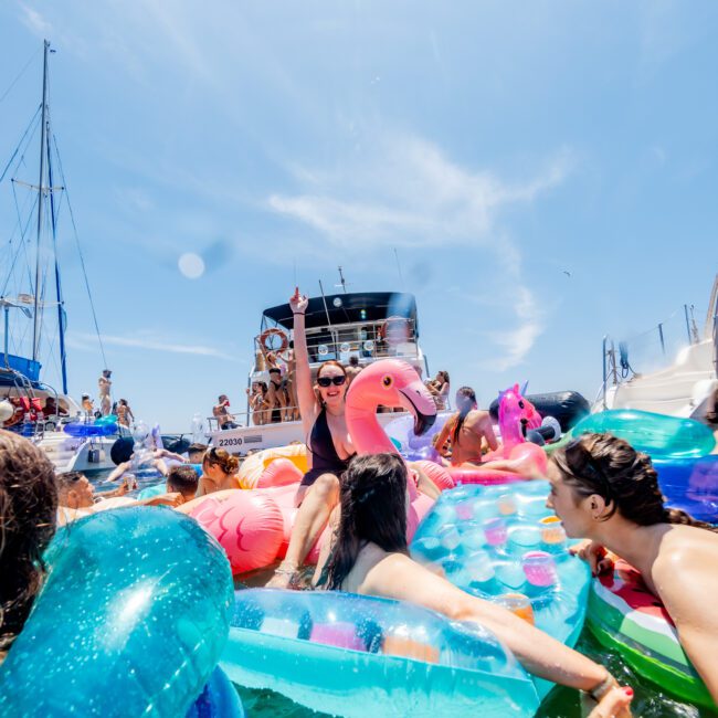 A lively scene of a boat party with people enjoying the water on colorful inflatable floats, including a pink flamingo. The sky is clear and blue. In the background, more people are on a boat. The atmosphere is festive and sunny.