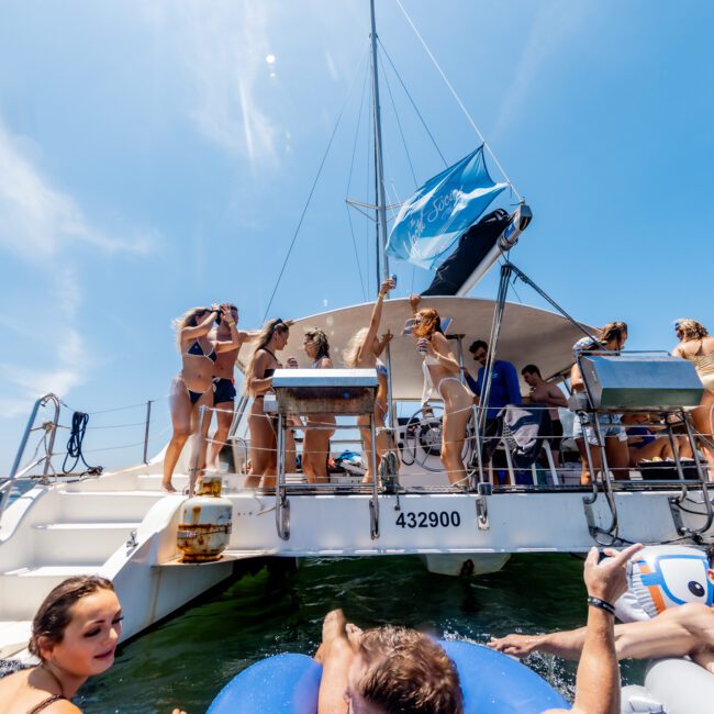 A group of people in swimwear are enjoying a sunny day on a boat. Some are standing, chatting, while others are in the water near the boat. A blue flag flies from the boat's mast. The sky is clear and blue.