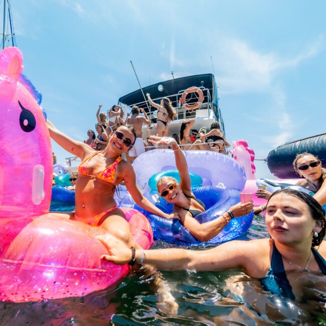 A group of people enjoying a sunny day on the water. They are swimming and relaxing on inflatable floaties, including a pink unicorn. A large boat is in the background. Smiles and laughter suggest a cheerful atmosphere.