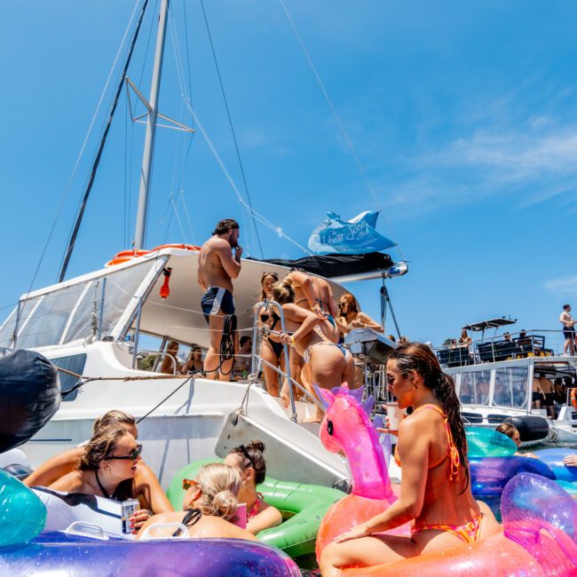 A group of people enjoy a sunny day on a boat and in the water. Some are on colorful inflatable floats, including a pink flamingo. The boat has a sail, and the sky is clear and blue. The atmosphere is lively and festive.