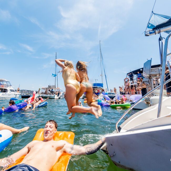 People enjoy a sunny day at a yacht party. Two women jump into the water from a boat. Others relax on floating inflatables and nearby boats. The scene is lively, with clear blue skies and sailboats in the background.