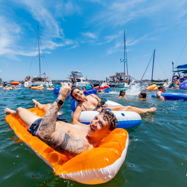 People are enjoying a sunny day on the water, floating on colorful inflatable rafts near several docked boats. The sky is clear and blue, and everyone appears relaxed and cheerful.