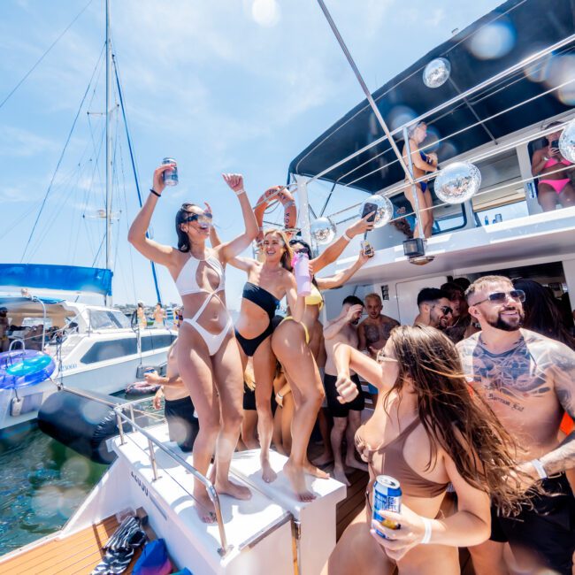 A group of people enjoying a sunny day on a boat. Three women in swimsuits dance on a platform while others, holding drinks, cheer around them. The background features a blue sky and another boat on the water.