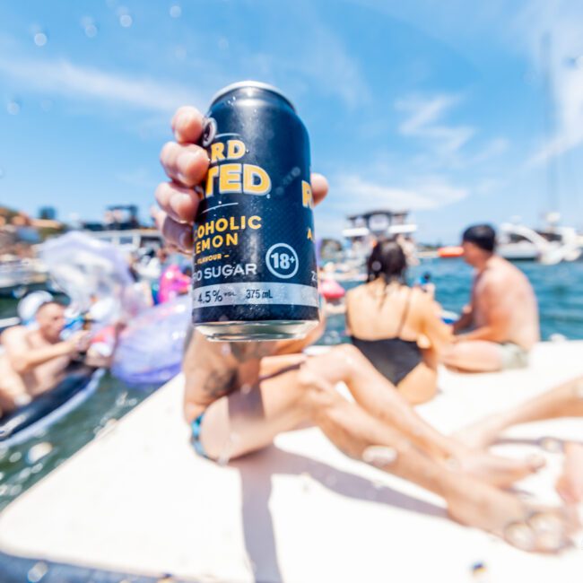 A person holds a can of hard seltzer with an 18+ label, 4.5% alcohol content, in front of a summer scene. People are sitting on a floating platform in the water, with boats and other swimmers in the background under a bright blue sky.