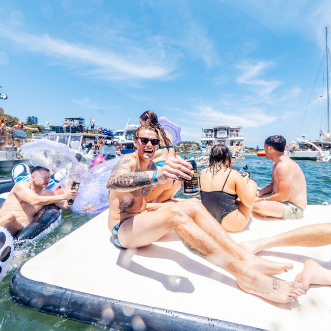 A group of people enjoying a sunny day on the water, sitting on a floating platform surrounded by boats. The central figure, holding a drink, appears to be laughing. Inflatable toys are visible, and others are swimming nearby.