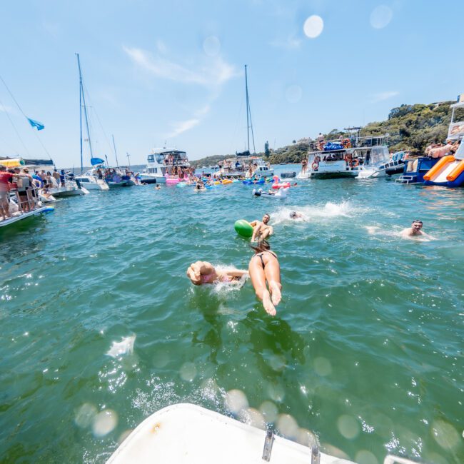 People enjoying a sunny day on a lake surrounded by boats. Some are swimming, and a person in a swimsuit is diving into the water. Floating toys and inflatables are visible, along with others on the boats. Lush hills are in the background.