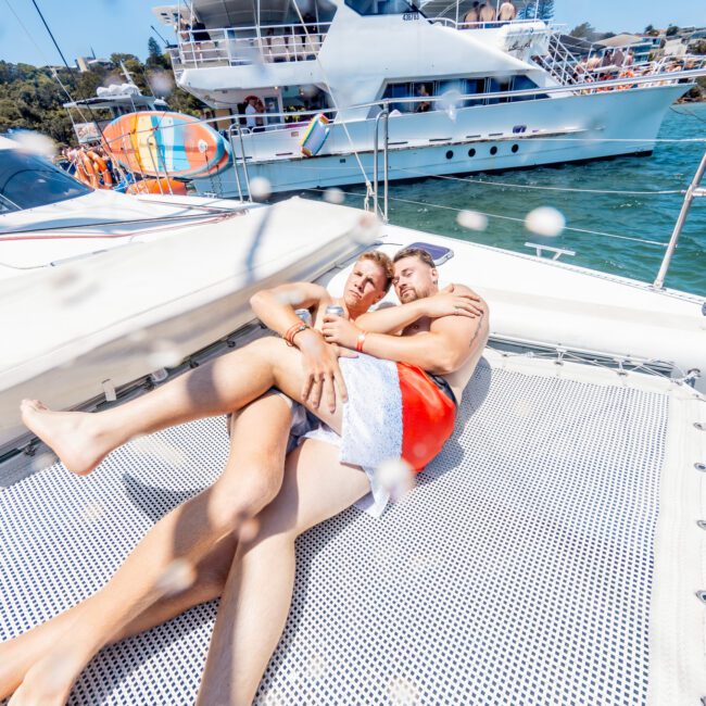 Two people are lying and embracing on the deck of a boat under a sunny sky. They are surrounded by water and other boats. The atmosphere appears relaxed and joyful.