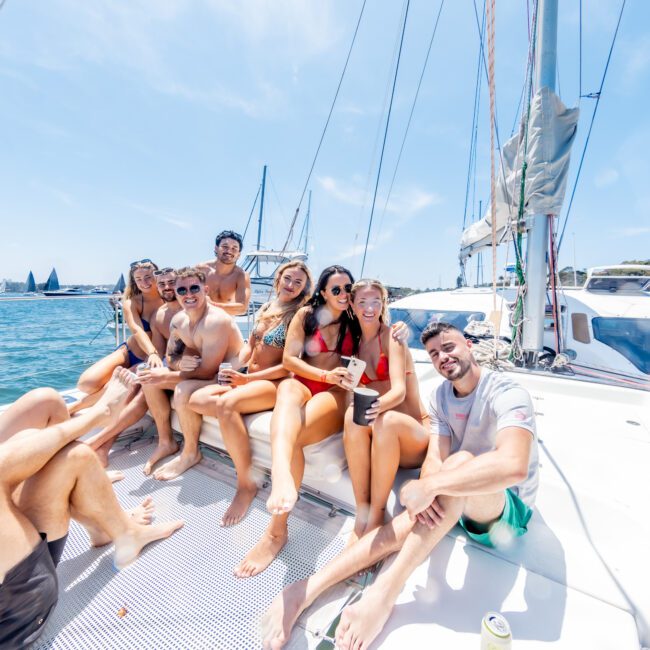 A group of people smiling and posing while sitting on the deck of a sailboat. It's a sunny day, and the ocean is calm. Some people wear sunglasses, and there's a drink visible on the deck. Other sailboats are in the background.