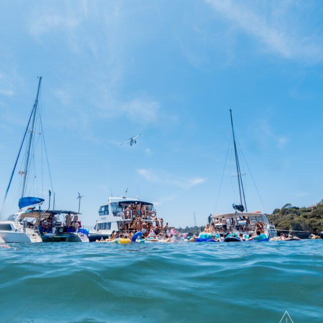 A group of people enjoying a party on several yachts and inflatables in the water under a clear blue sky. A small plane flies overhead. Lush greenery is visible in the background.