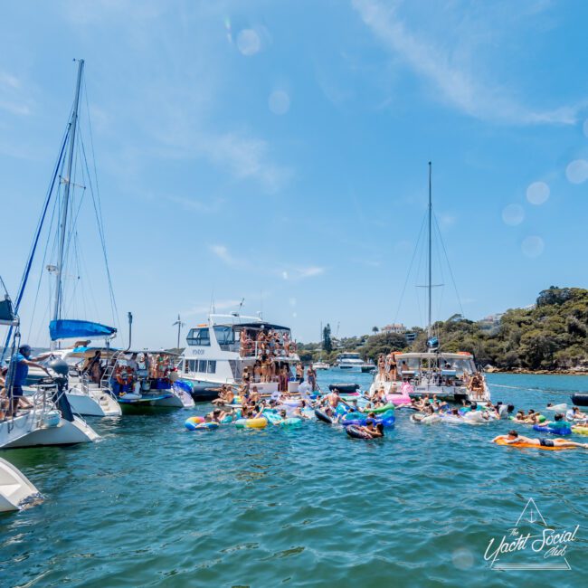 A lively gathering of people enjoying a sunny day on the water. Groups are floating on inflatables between several yachts and boats anchored nearby, with trees and a clear sky in the background.