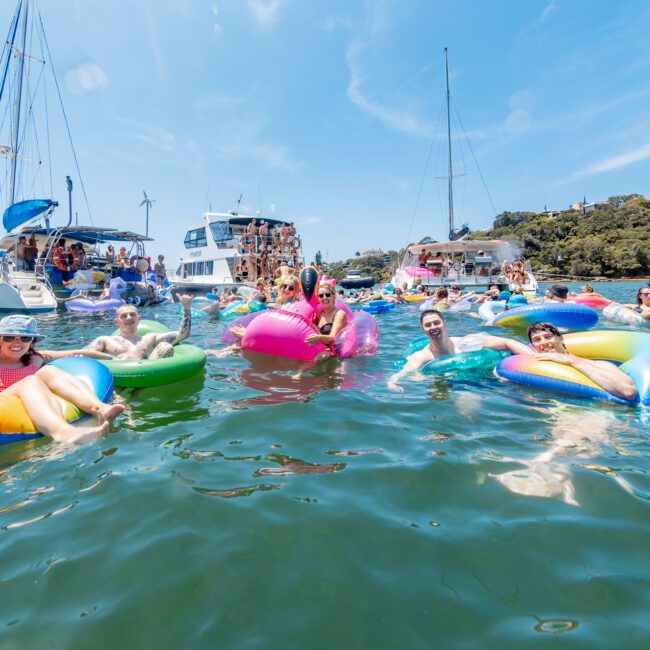 A group of people relax on colorful inflatable floats in the water, surrounded by boats. They are enjoying a sunny day, smiling and lounging. The scene is festive and lively, with clear skies and lush greenery in the background.