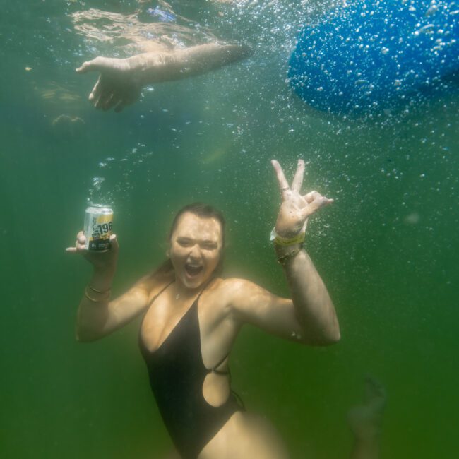 A person underwater in a black swimsuit holds a can and makes a peace sign. Bubbles and light create a playful atmosphere. A partially visible hand and object are seen above. Logo "The Yacht Social Club" is in the bottom corner.