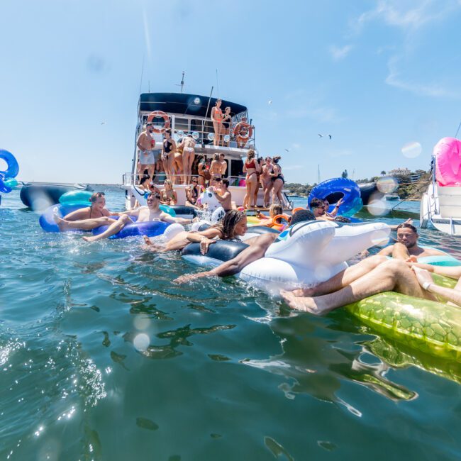 A lively scene of people enjoying a sunny day on the water. They are floating on colorful inflatables and socializing near anchored boats. The background shows a clear blue sky and scattered boats.