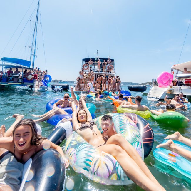 A group of people enjoying a sunny day on the water, floating on inflatable rafts and swim rings. In the background, a boat is filled with more people celebrating. The scene is lively and festive, with clear blue skies overhead.