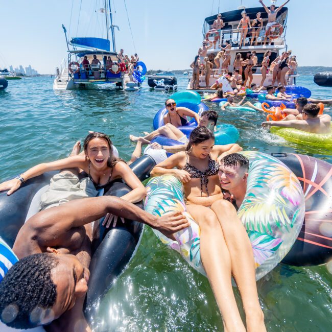 A lively group of people float on inflatables in a sunny water party near several boats. Everyone is smiling, enjoying the sunshine and water. The clear blue sky and distant city skyline add to the vibrant summer atmosphere.