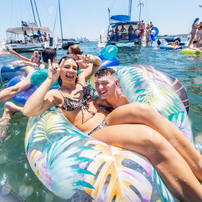 Two people smile and pose on a colorful inflatable float in the water, surrounded by others enjoying a boat party. Sunlight reflects off the water, while boats and more floats are visible in the background.