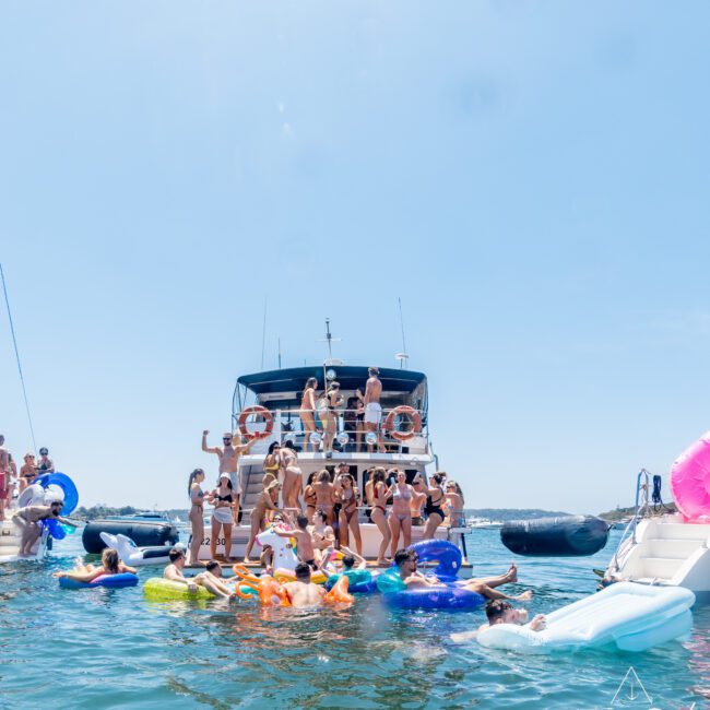 A group of people enjoy a sunny day on a large yacht, with many splashing and relaxing on colorful inflatables in the water. The sky is clear, and everyone appears to be having fun.