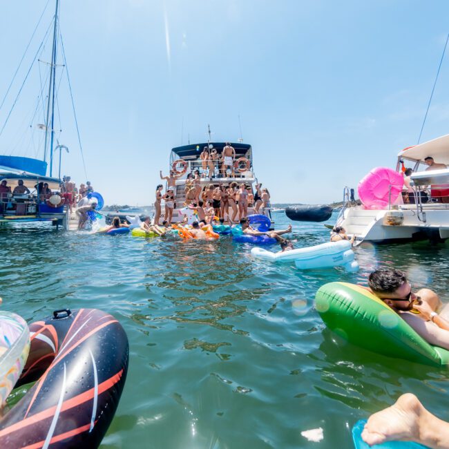 A lively scene of people enjoying a sunny day on the water. Several individuals are relaxing on colorful inflatable floats near anchored yachts, with more people visible on the boats. The sky is clear and bright.