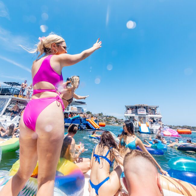 A crowd of people enjoying a sunny day on a lake, surrounded by inflatable rafts and boats. A woman in a pink swimsuit is standing and waving. The scene is lively and colorful with clear blue skies overhead.