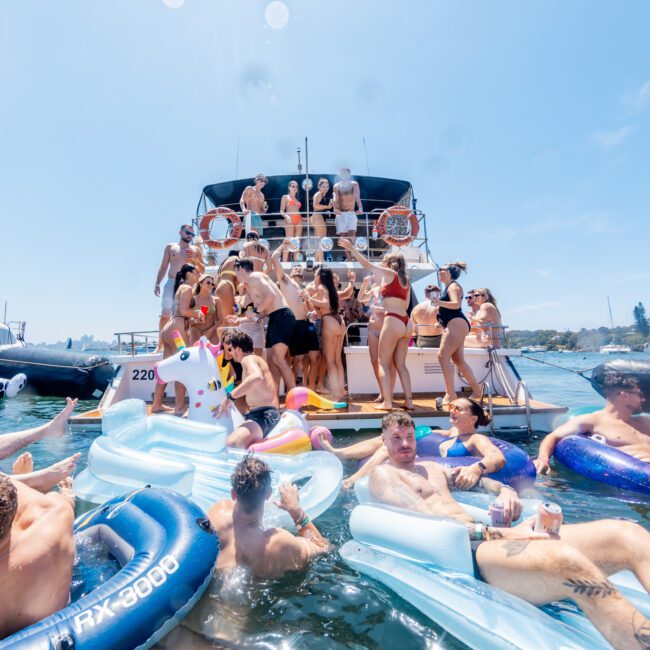 A lively gathering of people on a yacht and in the water on inflatable floats, enjoying a sunny day. The group seems to be socializing and having fun, surrounded by clear blue skies and water.