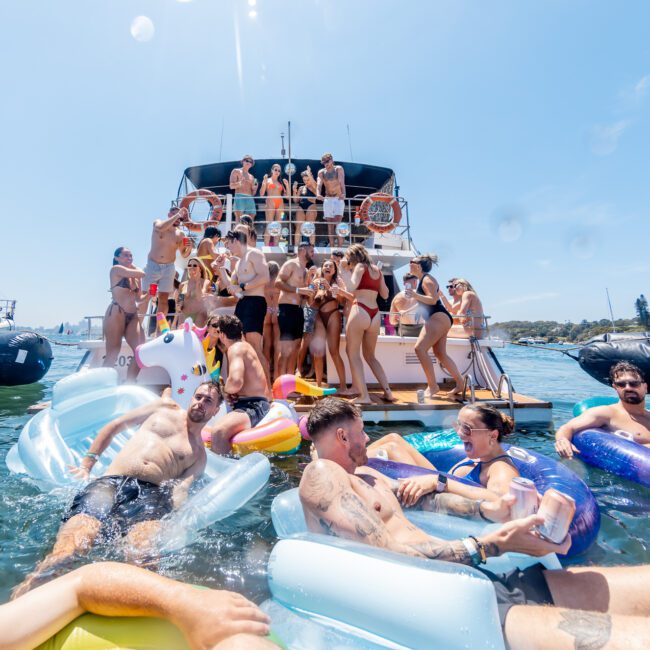 A vibrant scene with people enjoying a party on a boat and in the water. Some are on inflatable toys, including a unicorn float. The boat is crowded with partygoers under a clear blue sky.