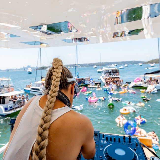 A DJ with long braided hair performs on a boat, overlooking a lively scene of boats and people on colorful inflatables in the water. The sky is clear and the atmosphere is festive.