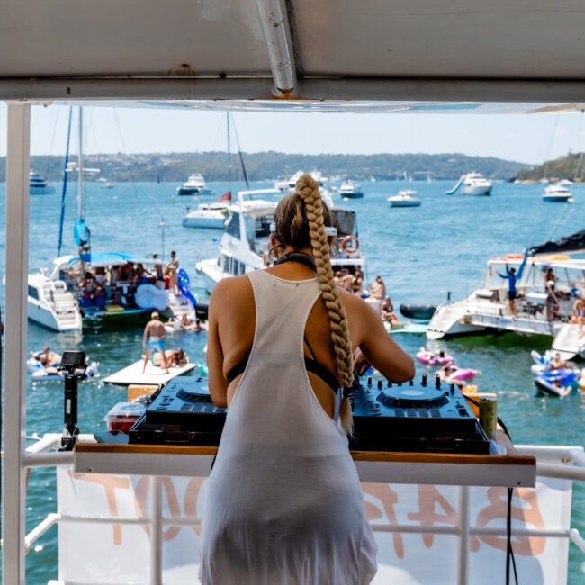 A DJ with long braids is performing on a boat deck, overlooking a lively scene of boats and people swimming in the sea. The clear blue water and sunny weather create a festive atmosphere.