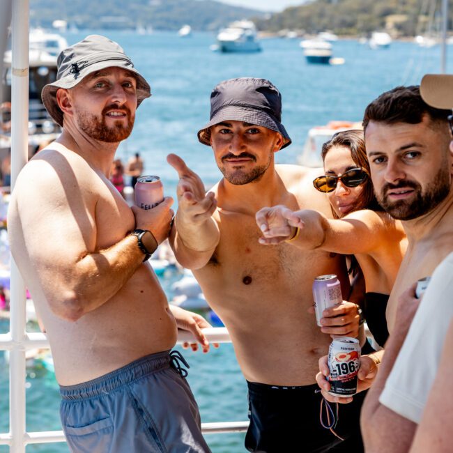 A group of four people on a boat, wearing swimwear and hats, enjoying a sunny day. They are holding drinks and smiling, with a scenic view of the water and distant boats in the background.