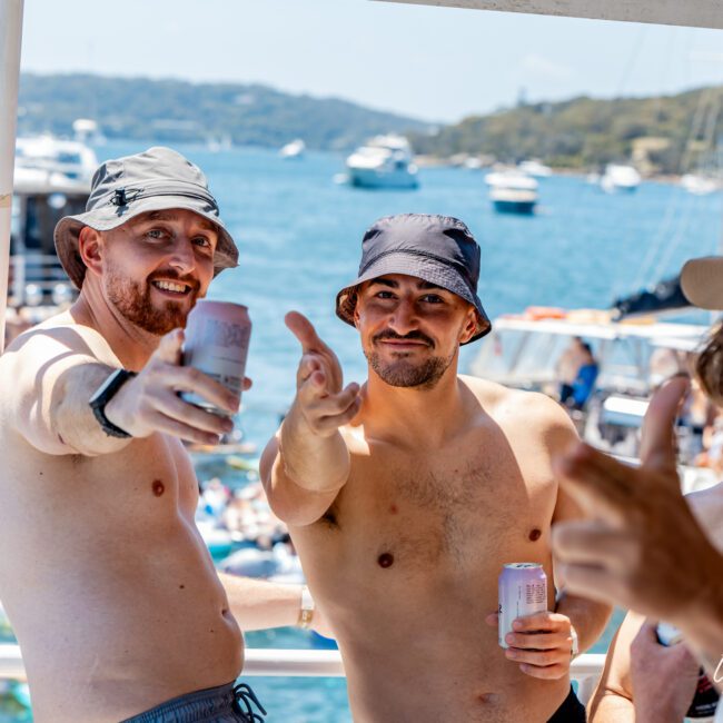 Three men in bucket hats and swimwear enjoy drinks on a boat. They're smiling and gesturing toward the camera, with a sunny ocean and distant boats in the background.