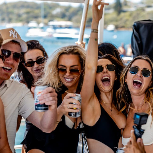 A group of friends enjoying a sunny day on a boat. They are smiling and holding drinks, with a scenic waterfront in the background. Most are wearing sunglasses and summer attire, capturing a lively and joyful moment on the water.