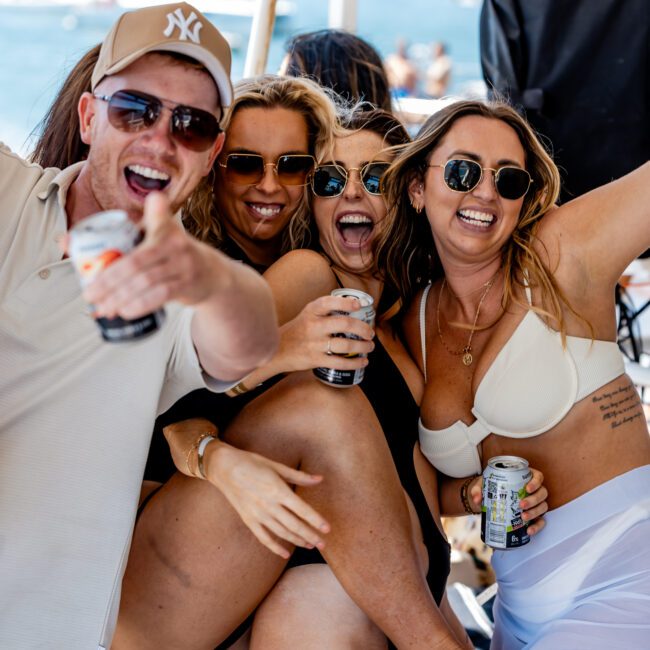 A group of four friends smiling and posing energetically on a boat. They are wearing sunglasses and swimwear, holding drinks, with a backdrop of water and anchored boats. A sunny day suggests a fun, social outing.