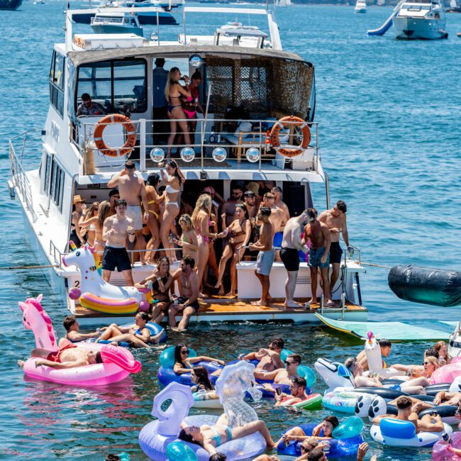 A crowded boat party on the ocean with people swimming around on colorful inflatable floats, including a unicorn and flamingo. The boat's deck is filled with more people enjoying the sunny day. Other boats and a distant hilly shoreline are visible.