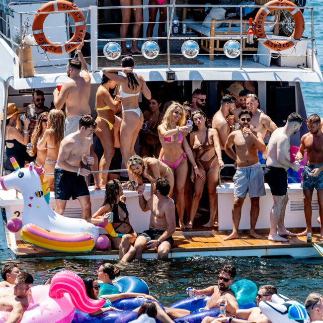 People are having a lively party on a boat, surrounded by colorful inflatables like unicorns and flamingos in the water. The group is enjoying the sunny day, some are dancing, while others relax and chat.