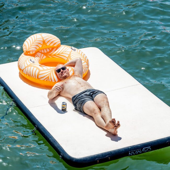 A person relaxes, lying on a floating mat in the water, wearing shorts and sunglasses. There's a tropical-themed inflatable beside them and a can of drink near their hand.