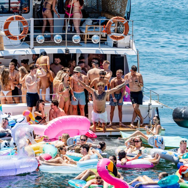 A group of people enjoying a party on a yacht and in the water, with colorful inflatable toys. Some are standing on the yacht, while others float around, all under a bright, sunny sky.