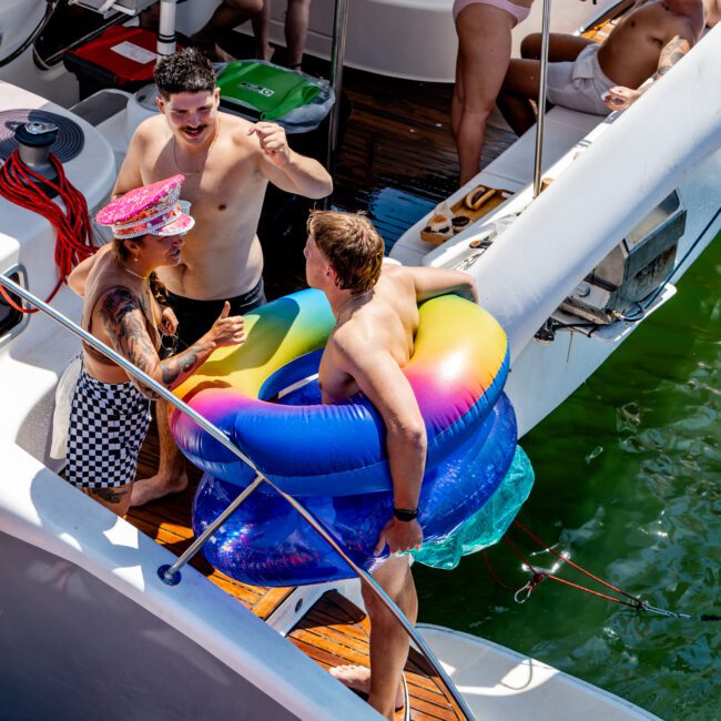 A group of people enjoying a sunny day on a boat. One person holds colorful inflatable rings, while others are socializing, including a person in a pink captain's hat and checkered outfit. The boat is docked on green water.