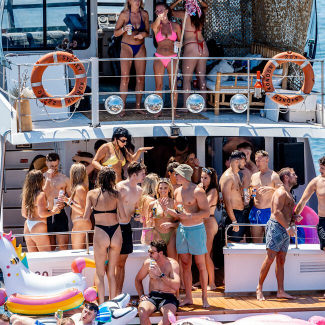 A large group of people in swimsuits partying on a boat deck and in the water, surrounded by inflatable floats. The scene is lively, with many people socializing under a clear blue sky.