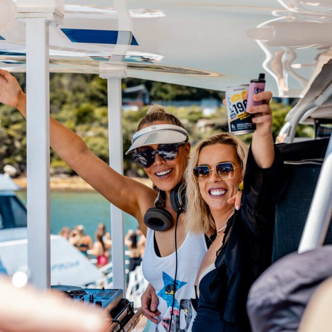 Two smiling women enjoying a sunny day on a boat. One wears headphones and a visor, while the other holds a drink can. Other boats and people are visible in the background.