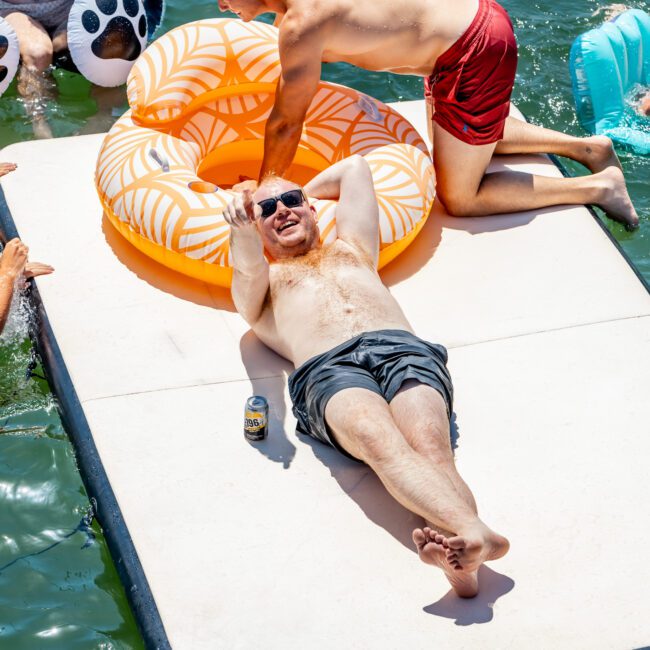 A man relaxes on a floating mat in the water, wearing sunglasses and gesturing with his hand. Another man nearby kneels, holding a large orange float. Other people are enjoying the water with inflatable toys around them.