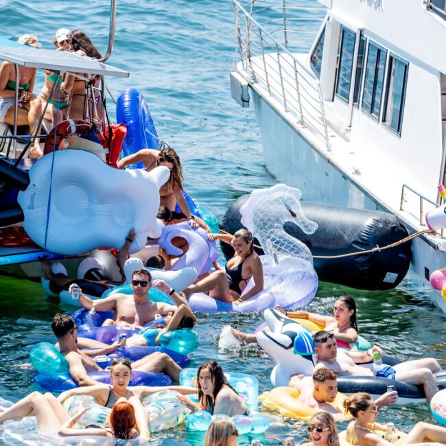 A vibrant scene of people enjoying a sunny day on the water, surrounded by boats. Many are relaxing on inflatable floats, including unicorns and swans, while others mingle and swim in the turquoise water. The atmosphere is lively and festive.