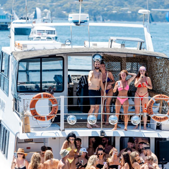 A group of people in swimwear enjoying a party on a boat. Some are dancing and socializing on the deck. The boat is on a large body of water with several other boats in the background. It appears to be a sunny day.