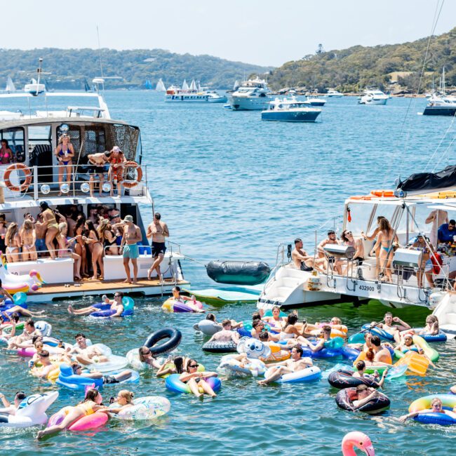 A lively scene of people enjoying a sunny day on a lake. Two boats are anchored with people socializing on deck and in the water, using colorful inflatables. The background shows more boats and a tree-lined shoreline.