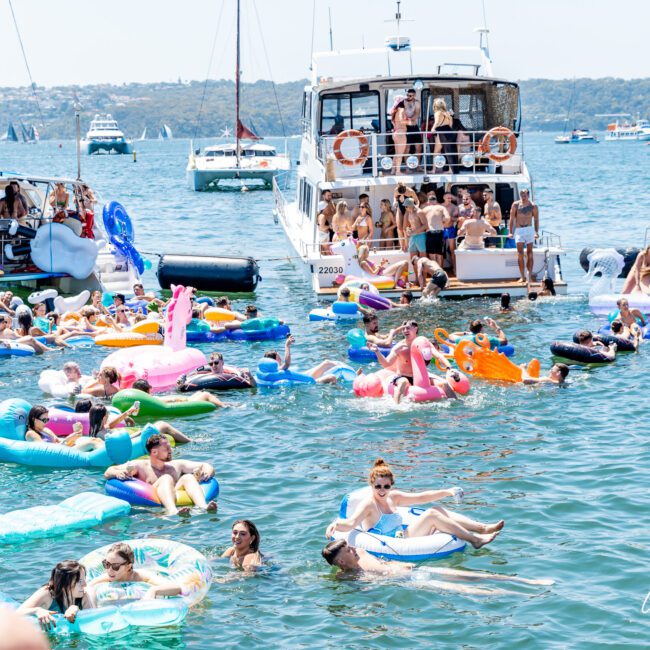 People are enjoying a sunny day on a lake, surrounded by boats. Many are on colorful inflatable rafts and pool floats. The scene is lively, with groups socializing and the water filled with various floats shaped like animals and objects.