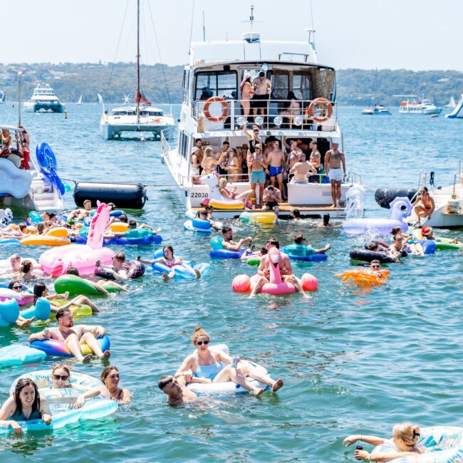 A lively scene of people enjoying a sunny day on a lake. Many are floating on colorful inflatables near a large yacht. The water is dotted with various inflatable toys, and more boats are visible in the background.