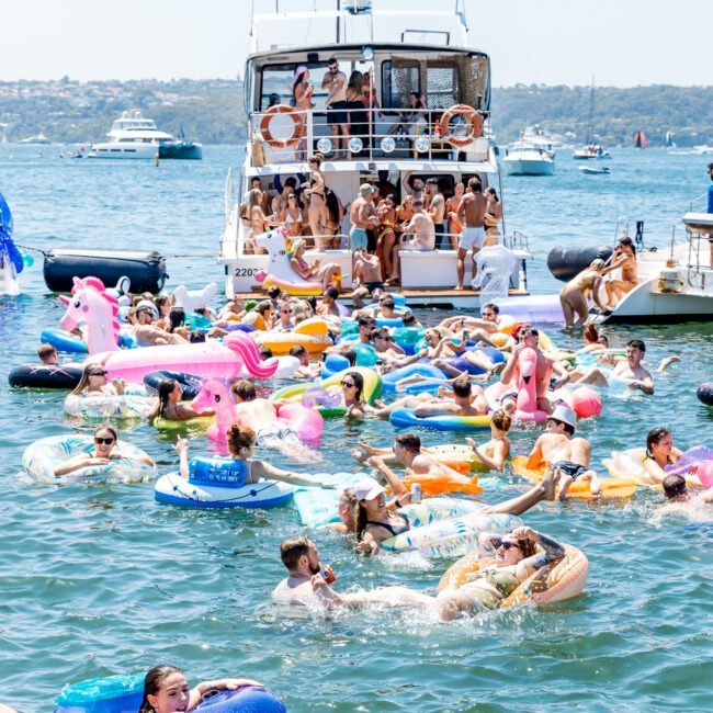 A lively scene of people enjoying a sunny day on the water, surrounded by colorful inflatables, such as unicorns and flamingos. Several boats are anchored nearby, and the shoreline is visible in the distance under a clear blue sky.